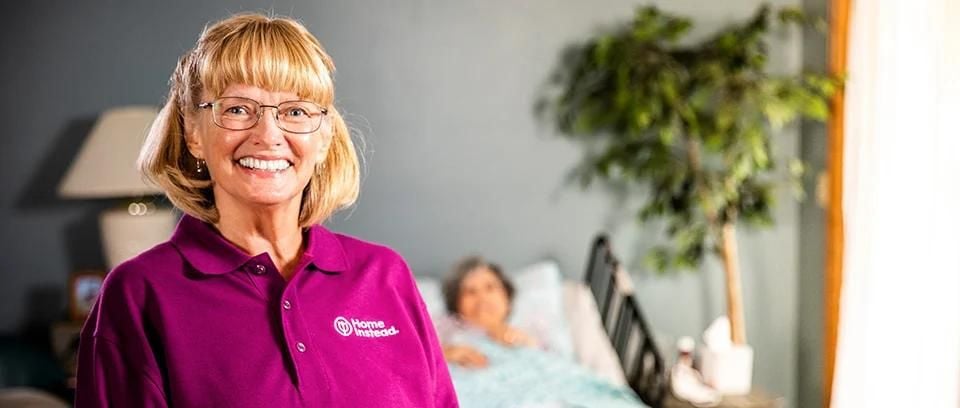 smiling caregiver with senior client in bed in the background
