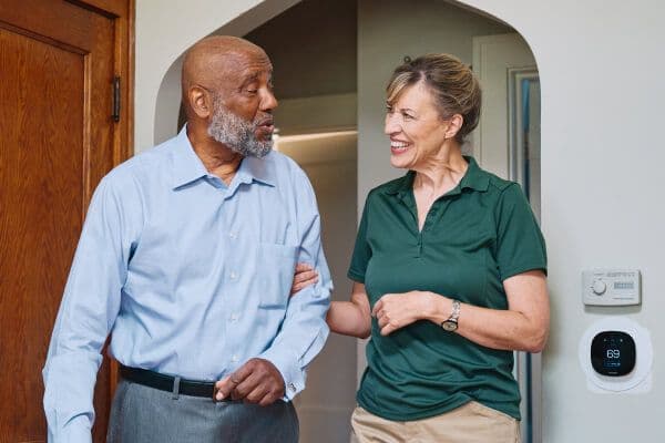 A Home Instead caregiver and an elderly man walk arm-in-arm through a doorway, both smiling, symbolizing trust and companionship in daily activities.