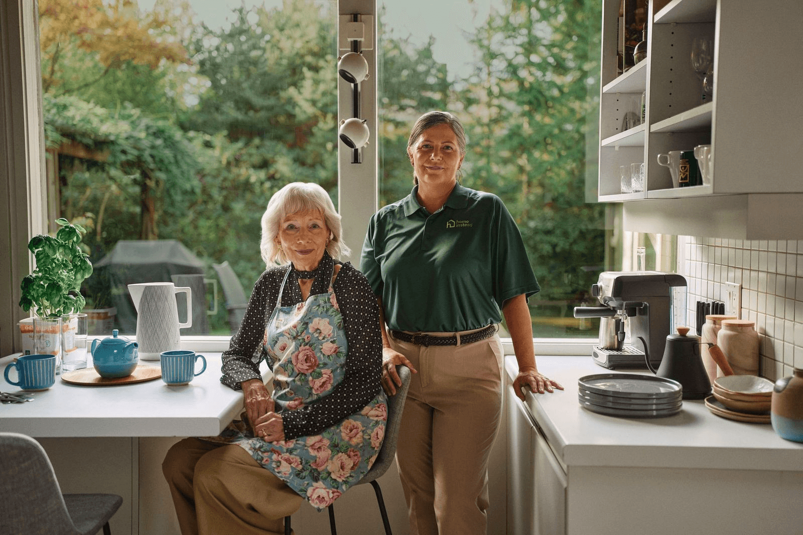 An adult daughter with her arm around her smiling mother in a kitchen
