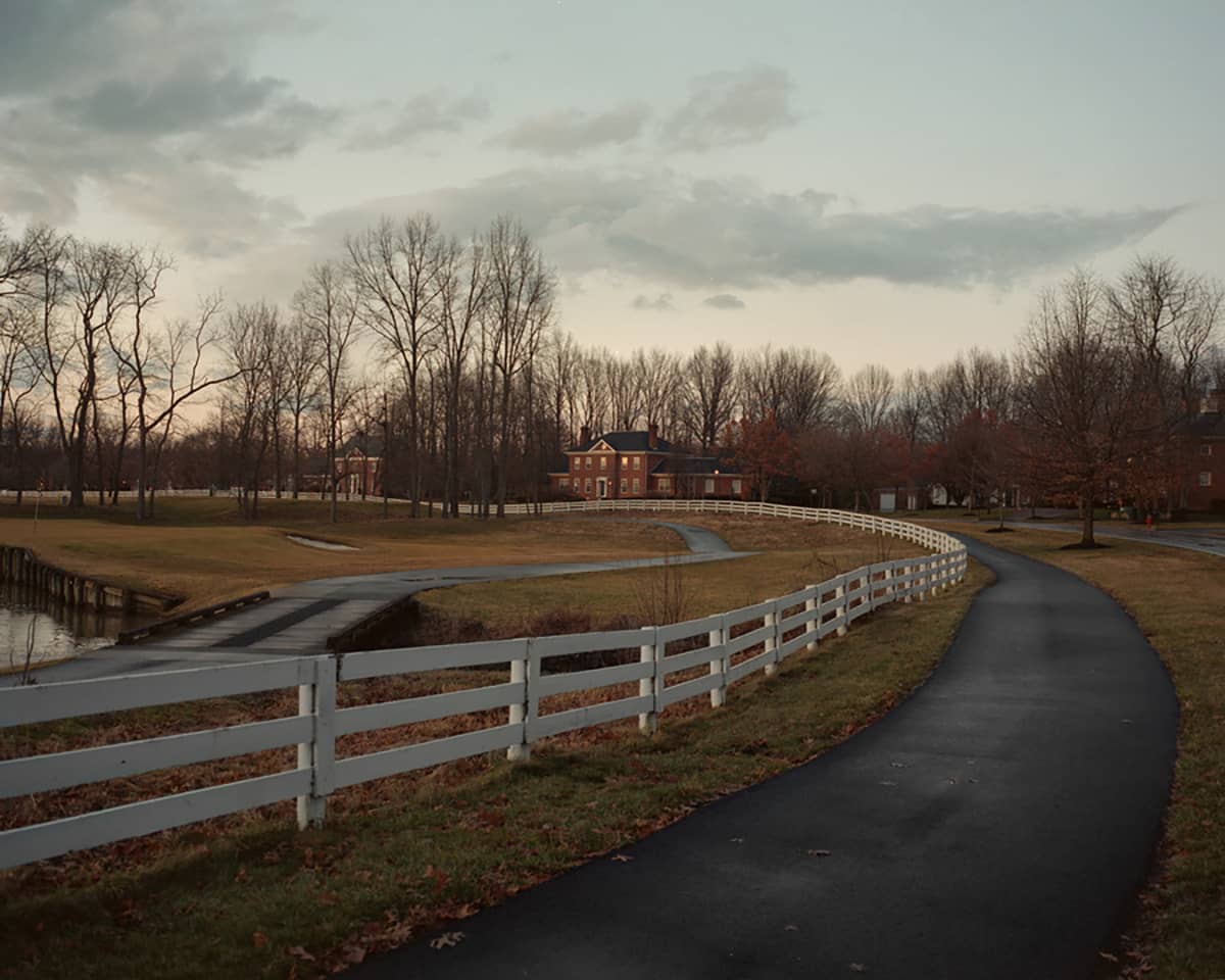 Photo of white fence around a putting green along a driveway leading up to a farm house.
