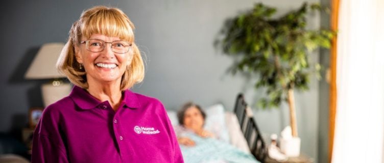 smiling home instead caregiver with senior in background laying in bed