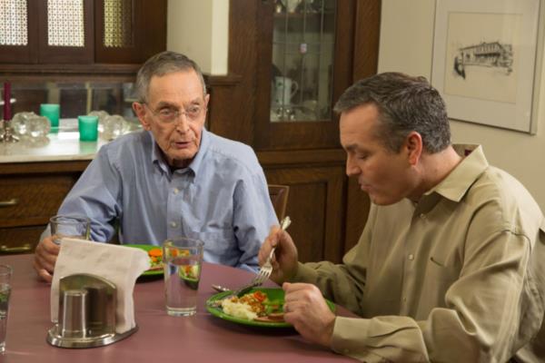 Senior man and son eat and talk at kitchen table at home.