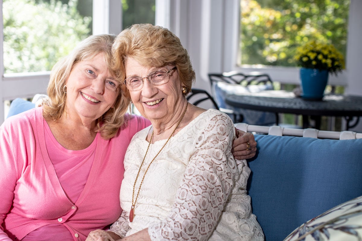 Home Instead Caregiver and senior woman smile and hug while sitting on porch.