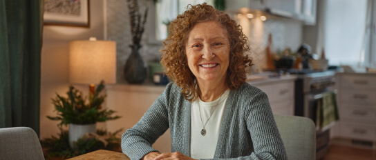 An elderly woman sits at a kitchen table, smiling warmly, with holiday decorations in the background, representing comfort and connection.