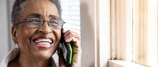 A joyful elderly woman talks on a cell phone by a sunny window, showcasing connection and communication as she calls her local Home Instead.