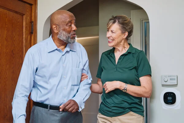 A Home Instead caregiver and an elderly man walk arm-in-arm through a doorway, both smiling, symbolizing trust and companionship in daily activities.
