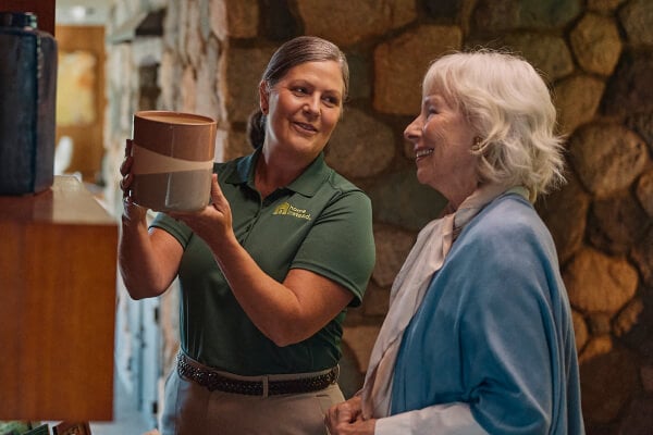 A Home Instead caregiver in a green uniform helps an elderly woman examine a piece of pottery in a warmly lit home, symbolizing companionship and support with daily activities.