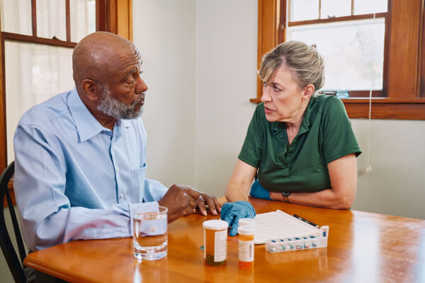 A Home Instead caregiver and an elderly man sit at a table discussing medications, with pill organizers and water, showcasing careful health management.