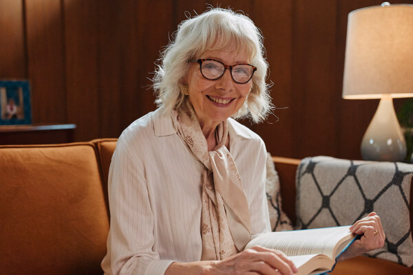 A smiling elderly woman wearing glasses sits on a cozy couch with a book in her hands, under warm lighting, highlighting comfort and independence.