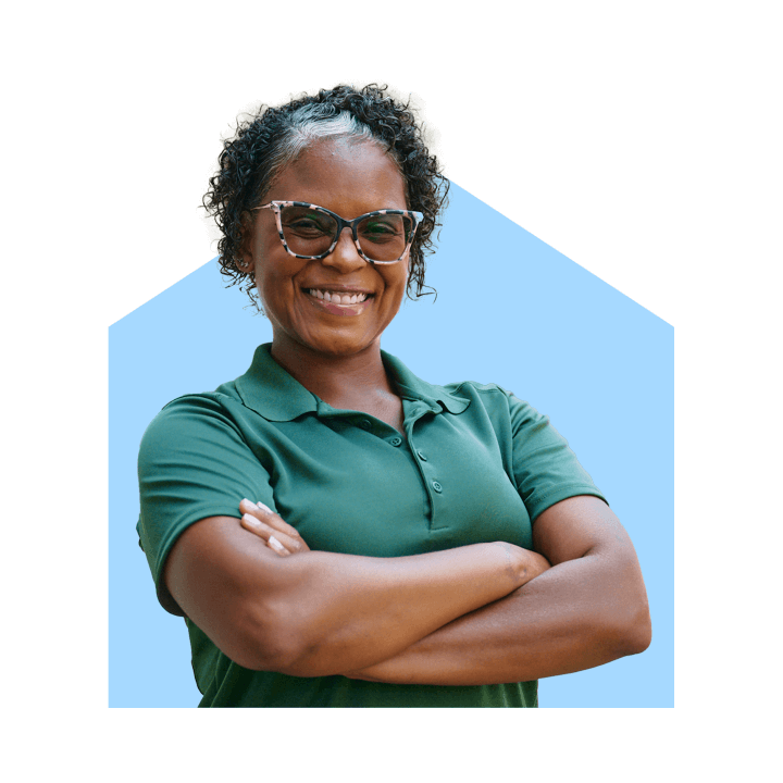 A confident Home Instead caregiver in a green uniform smiles with arms crossed, standing outdoors, representing trust and professionalism.