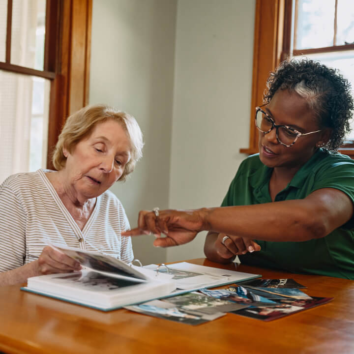 A Home Instead caregiver and an elderly woman review a photo album together at a dining table, sharing memories and stories.