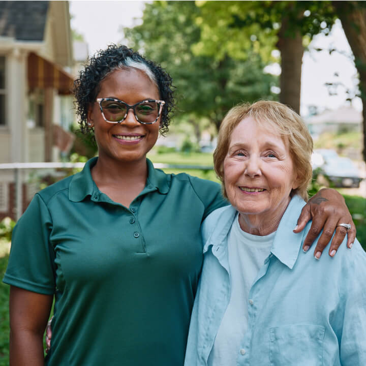 A Home Instead caregiver with an arm around an elderly woman outdoors, both smiling warmly, showcasing trust and companionship.