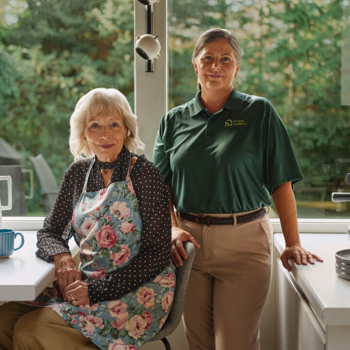 A Home Instead caregiver in a green uniform stands beside an elderly woman in a floral apron in a bright kitchen, symbolizing meal prep, support and collaboration.