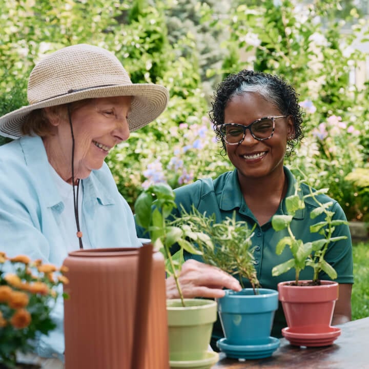 A cheerful elderly woman wearing a sun hat gardens alongside her Home Instead caregiver in a colorful and vibrant outdoor setting.