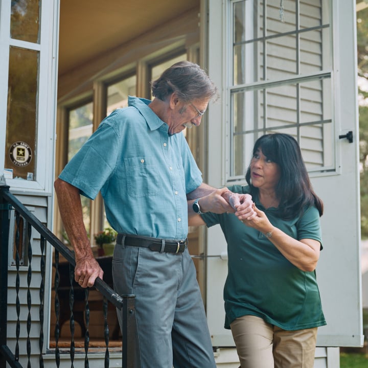 A Home Instead caregiver helps an elderly man, a veteran, safely walk down the front steps of a home, emphasizing Home Instead's care and thoughtful support.