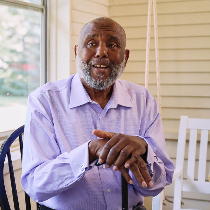 An elderly man with a beard sits on a sunlit porch in a light blue shirt, speaking animatedly and enjoying a peaceful moment outdoors.