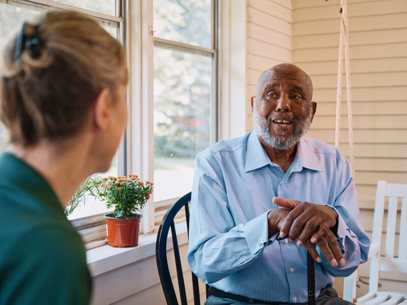 An elderly man gestures while speaking with his Home Instead caregiver on a bright porch, fostering a moment of friendly conversation and companionship.