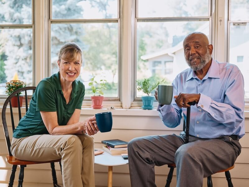 A Home Instead caregiver and an elderly man enjoy coffee together on a porch, showcasing shared moments of relaxation and connection.