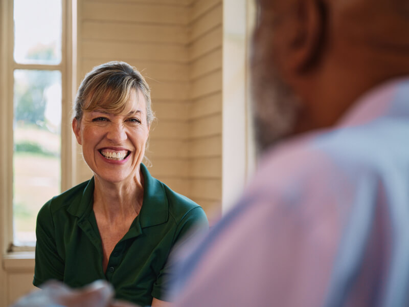 A Home Instead caregiver attentively listens to an elderly man as they chat in a sunlit room, highlighting meaningful connection and understanding.