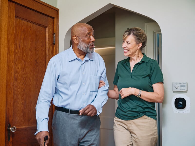 A Home Instead caregiver and an elderly man walk arm-in-arm through a doorway, both smiling, symbolizing trust and companionship in daily activities.