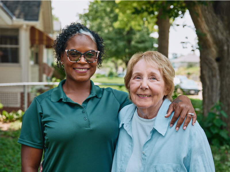 A Home Instead caregiver with an arm around an elderly woman outdoors, both smiling warmly, showcasing trust and companionship.