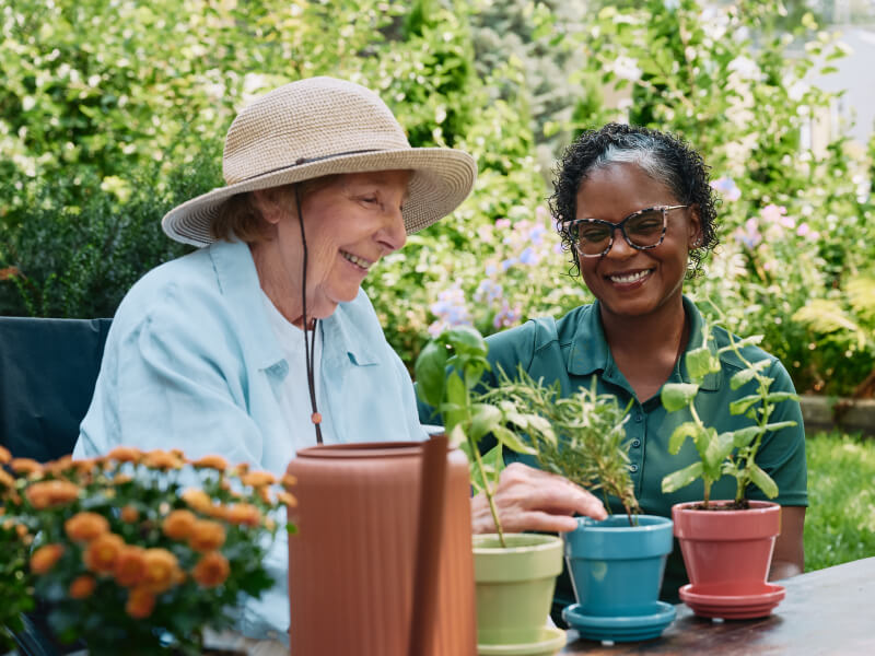 A cheerful elderly woman wearing a sun hat gardens alongside her Home Instead caregiver in a colorful and vibrant outdoor setting.