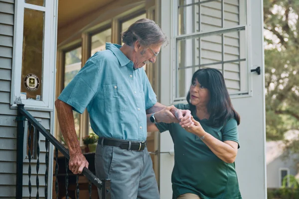 A Home Instead caregiver helps an elderly man, a veteran, safely walk down the front steps of a home, emphasizing Home Instead's care and thoughtful support.