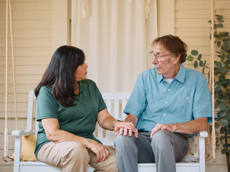 A Home Instead caregiver and an elderly man hold hands on a porch swing, fostering a sense of trust, reassurance, and emotional support.