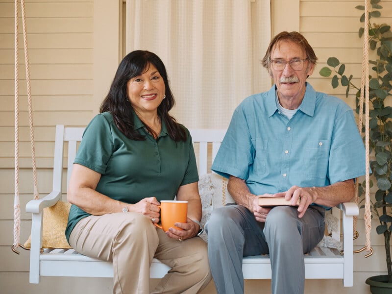 A Home Instead caregiver and an elderly man sit together on a porch swing, enjoying coffee and a book, emphasizing relaxed and meaningful companionship.
