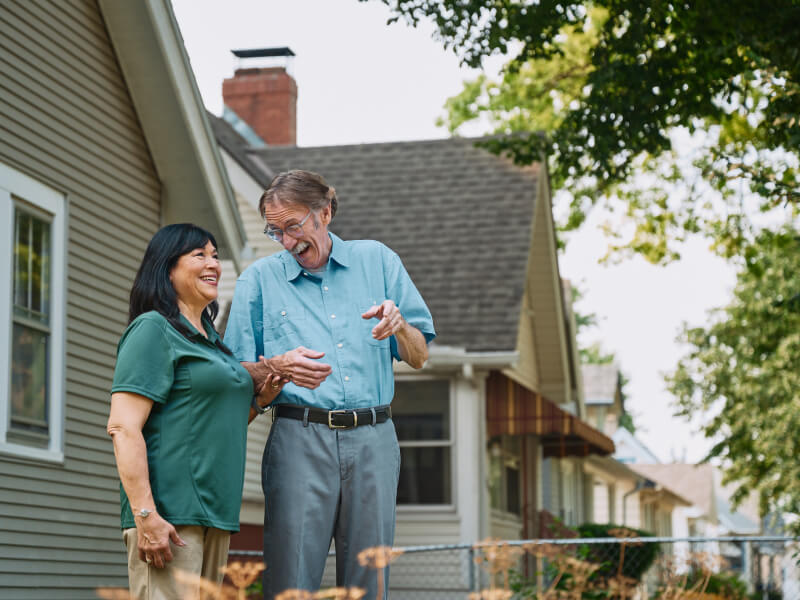 A Home Instead caregiver walks outdoors with an elderly man, both laughing and enjoying the fresh air, representing active engagement and companionship.