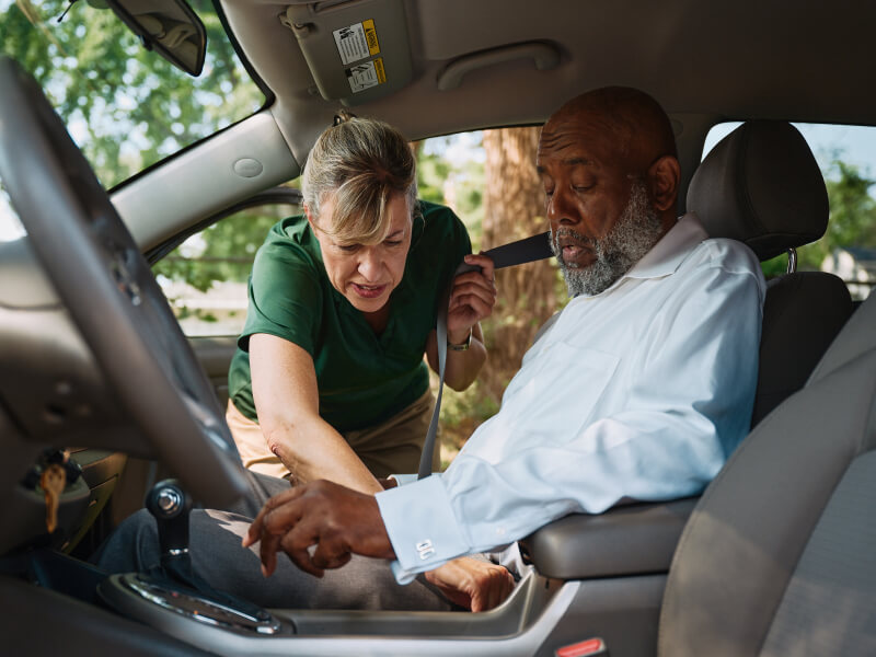 A Home Instead caregiver assists an elderly man in the passenger seat by fastening his seatbelt, ensuring safety and comfort during transportation.