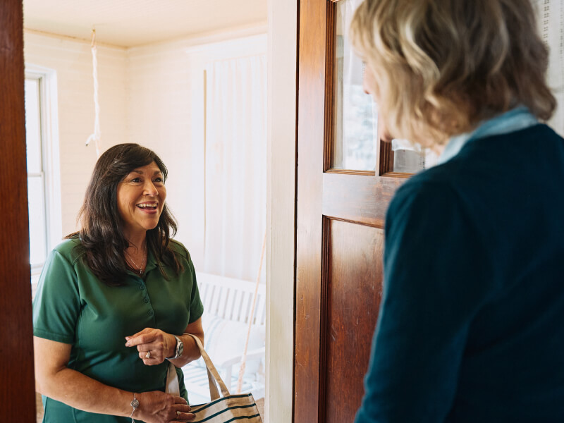 A Home Instead caregiver greets an elderly woman at the door with a warm smile, holding a tote bag, symbolizing friendliness and readiness to assist.