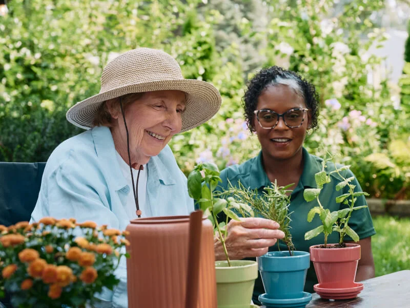 A Home Instead caregiver and an elderly woman smile together as they tend to potted plants outdoors, emphasizing companionship and shared hobbies.