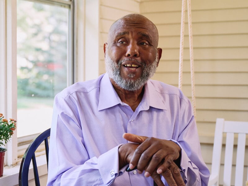 An elderly man with a beard sits on a sunlit porch in a light blue shirt, speaking animatedly and enjoying a peaceful moment outdoors.
