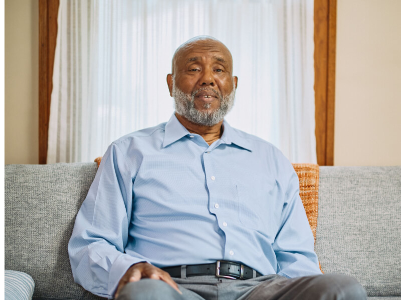 The same elderly man with a beard sitting on a comfortable sofa indoors, wearing a light blue shirt and appearing relaxed and at ease.