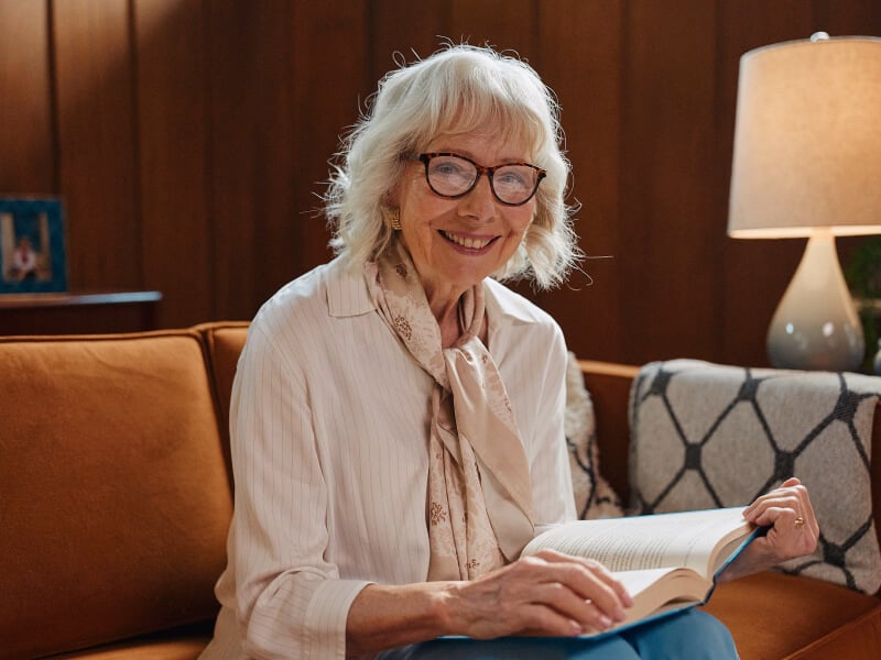 A smiling elderly woman wearing glasses sits on a cozy couch with a book in her hands, under warm lighting, highlighting comfort and independence.