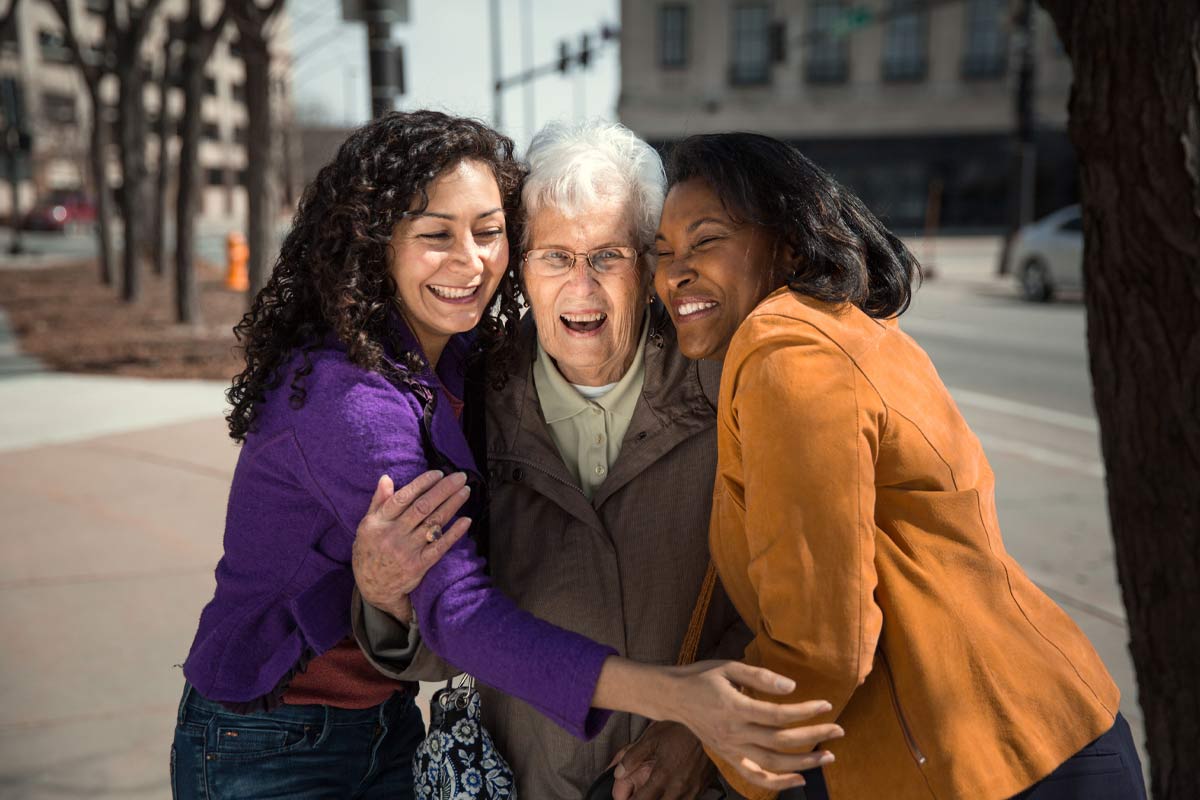 two female caregivers happily hug senior woman on a sidewalk