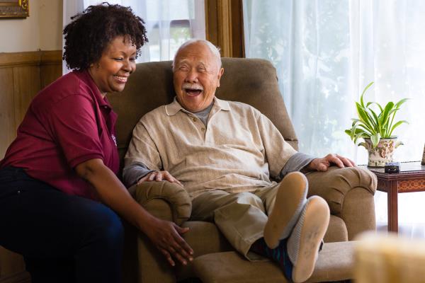 Home Instead Caregiver laughs with elderly man sitting in recliner at home.