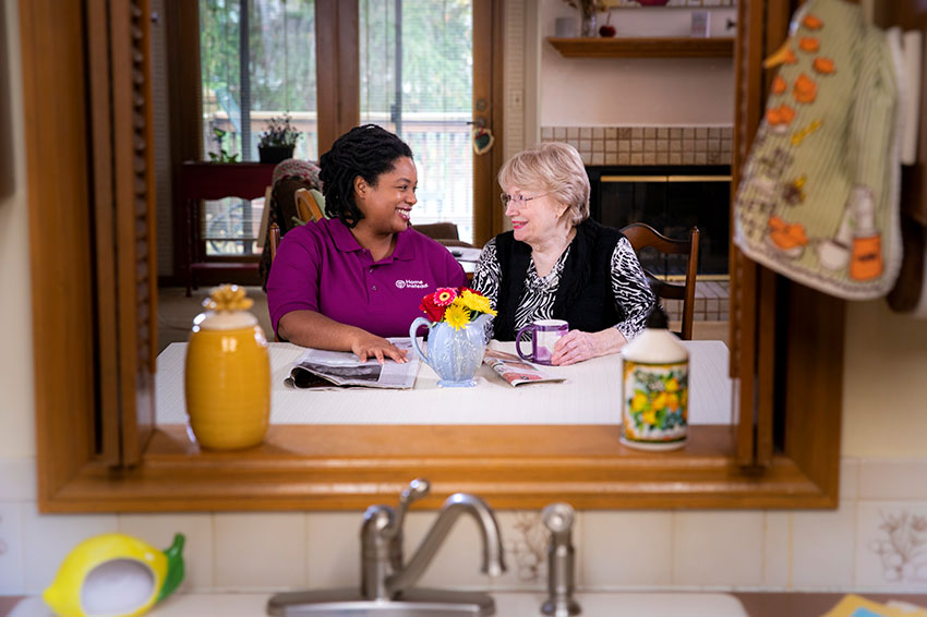 Home Instead CAREGiver and senior smiling at eachother sitting in kitchen