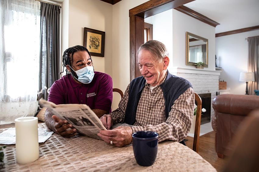 Home Instead Caregiver wearing mask reading newspaper with senior man at home