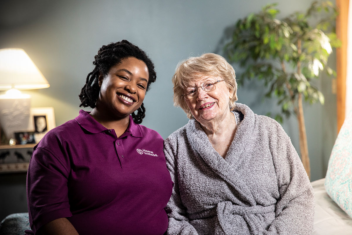 Home Instead Caregiver and senior woman smile while sitting together on bed at home