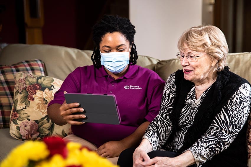 Home Instead Caregiver wearing mask helps senior woman with video conference call at home using tablet